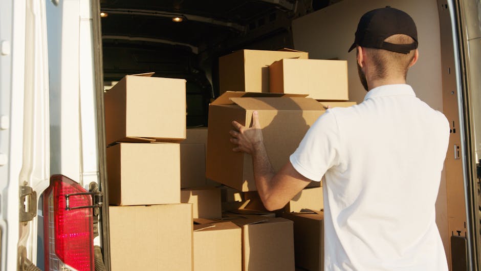 A man with a beard and wearing a black cap, a white polo shirt, and blue jeans, stands outside the open back doors of a white moving van. Inside the van, numerous brown cardboard boxes of various size